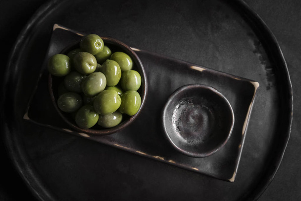 Aceitunas verdes en un recipiente de cerámica negra sobre un fondo de madera. Aceitunas servidas en un restaurante
 - Foto, Imagen