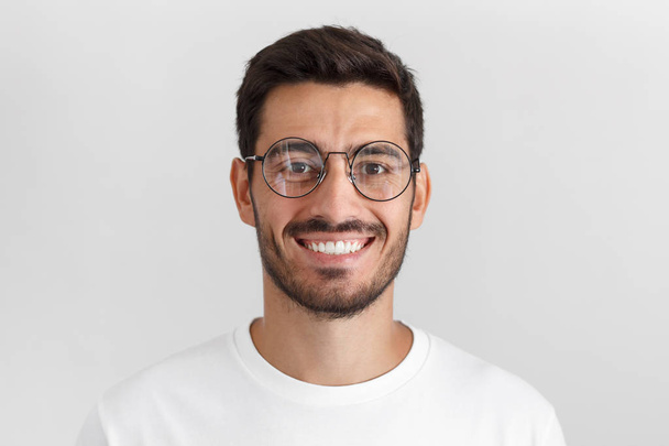 Daylight portrait of young handsome caucasian man isolated on grey background, dressed in white t-shirt and round eyeglasses, looking at camera and smiling positively - Фото, зображення