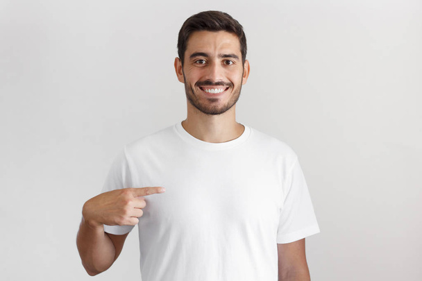 Daylight shot of smiling young man pointing at his blank white t-shirt with index finger, copy space for your ads, isolated on gray background - Foto, Bild
