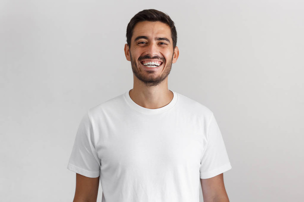 Horizontal shot of young positive european man isolated on gray background standing in blank white casual t-shirt looking straight at camera with happy smile, laughing - Foto, imagen
