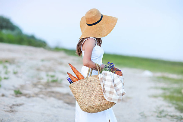 Молода жінка тримає кошик для пікніка з пляшкою вина і багет на піщаному пляжі
 - Фото, зображення