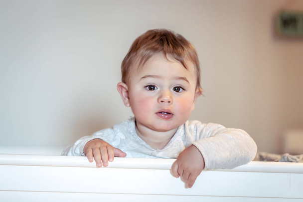 Portrait of a cute little boy standing in the crib in kids bedroom, pretty adorable baby wearing pajamas, happy healthy childhoo - Photo, Image
