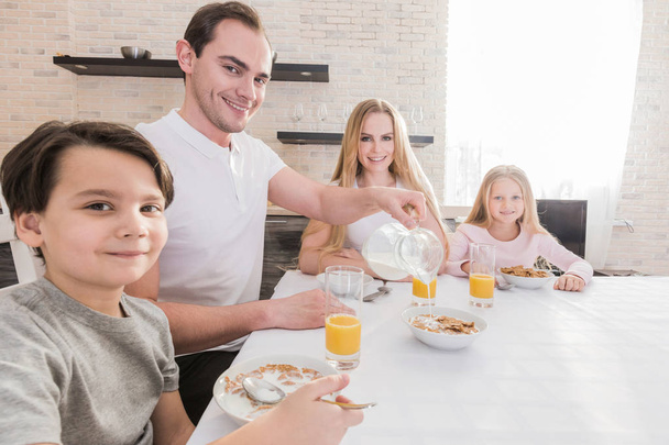 Szczęśliwych młodych rodziców i ich dzieci siedzą razem przy stole w kuchni, jeść płatki, mieć zdrowe śniadanie, cieszyć się dzień dobry, mieć przyjazne stosunki. Rodziny i jedzenie koncepcja. - Zdjęcie, obraz