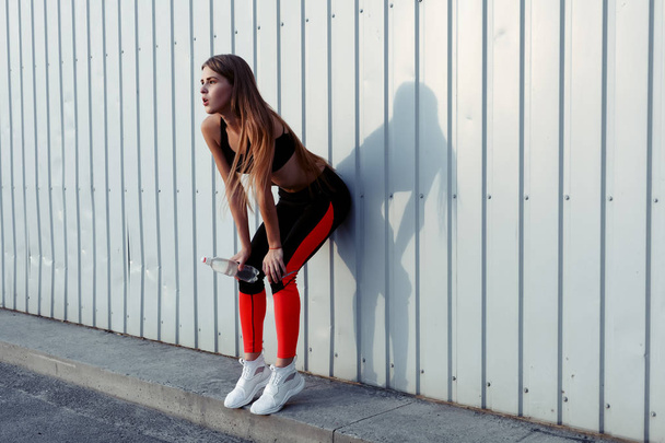 Female athlete drinking water while standing by a grey wall. Slim woman in sportswear taking a break after workout. - Foto, imagen