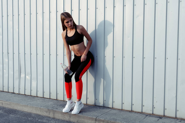 Female athlete drinking water while standing by a grey wall. Slim woman in sportswear taking a break after workout. - Foto, immagini