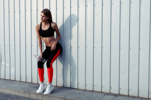 Female athlete drinking water while standing by a grey wall. Slim woman in sportswear taking a break after workout. - 写真・画像