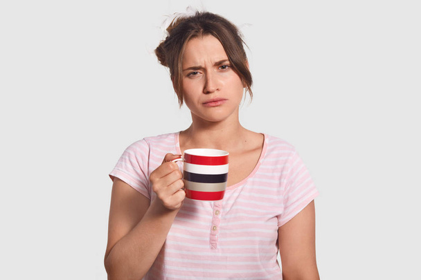 Upset sleepy lovely young woman holds cup of coffee, drink refreshing beverage to feel brave early in morning, dressed in casual clohes, has displeased expression, isolated over white background. - Фото, зображення