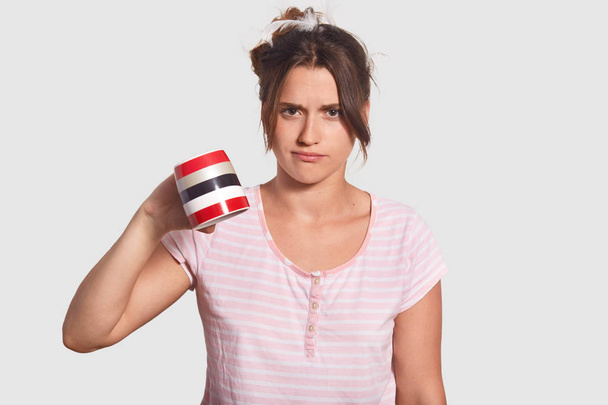Discontent upset woman in pyjamas keeps mug down, demonstrates that she has no coffee to drink, needs to go in shop, stands against white background. People, morning drink and awakening concept - Fotografie, Obrázek