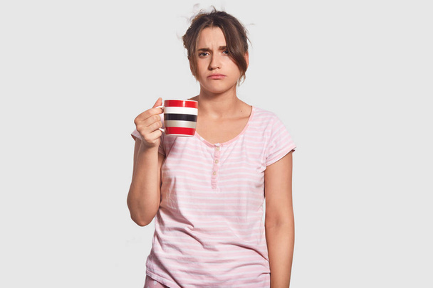Waist up portrait of unhappy woman has gloomy sleepy expression, tries to wake up, drinks hot coffee, doesnt want to go for work, dressed in domestic clothing, isolated over white background - Photo, image