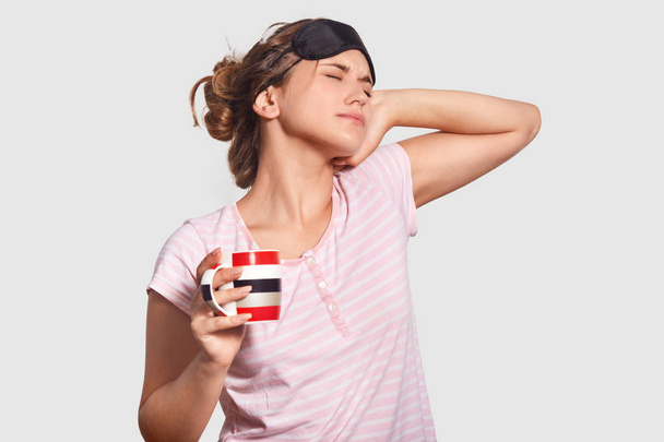 People and awakening concept. Sleepy young European female wears eye mask on head, pyjamas, stretches after getting up early, drinks hot coffee, cant open eyes, isolated over white background. - Foto, Bild