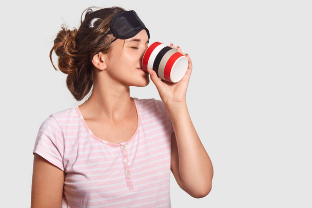 Young pretty female wakes up in morning, wears sleep mask and pyjamas, drinks hot coffee, tries to feel brave, isolated over white background. Woman enjoys refreshing drink after night rest. - Photo, image