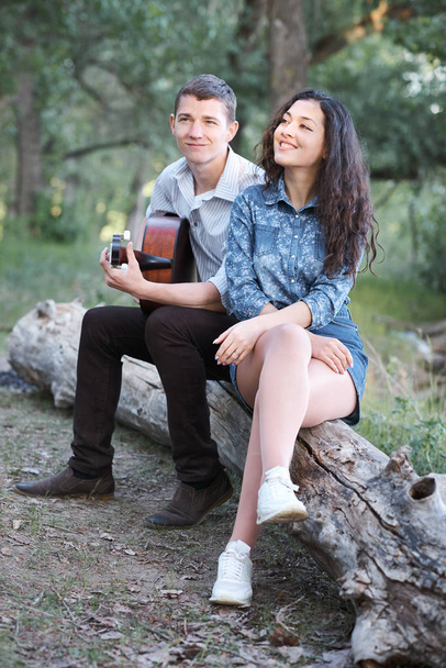 young couple sitting on a log in the forest and playing guitar, summer nature, romantic feelings - Foto, afbeelding