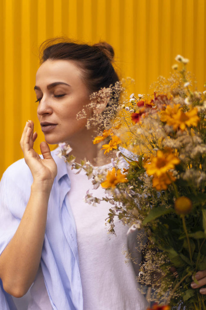 Hermosa mujer morena sobre fondo amarillo con moño en la cabeza con camisa azul y camiseta blanca mientras sostiene los campos de flores en su mano. Mujer trayendo a la flor
 - Foto, imagen