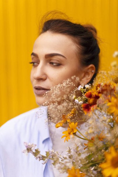 Beautiful brunette woman on yellow background with bun on her head wearing blue shirt and white t-shirt while holding fields flowers on her hand. Female bringing to flower - Foto, Bild
