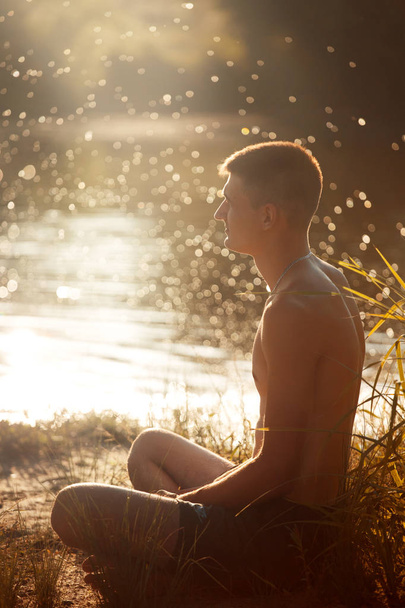Nuori mies, jolla on alaston vartalo mustissa shortseissa ja joka istuu joen rannalla kirjan ja medetiruetin kanssa auringonlaskussa.
. - Valokuva, kuva