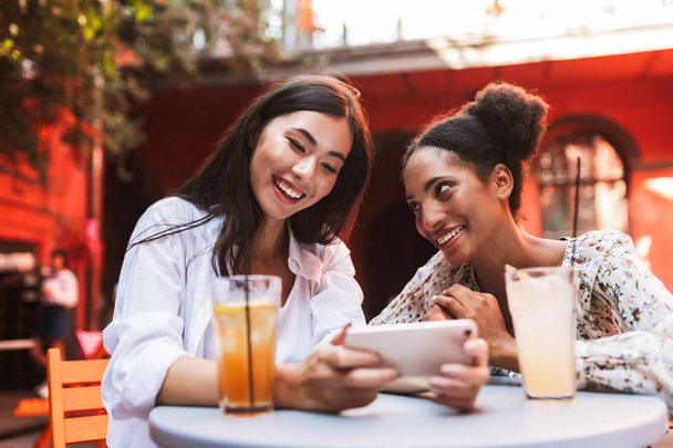 Pretty smiling ladies joyfully using cellphone together with drinks near on table spending time in courtyard of cafe - Foto, Imagen