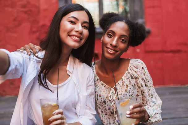 hübsche lächelnde Mädchen mit Cocktails in der Hand, die fröhlich zusammen Fotos machen und Zeit im Innenhof des Cafés verbringen - Foto, Bild