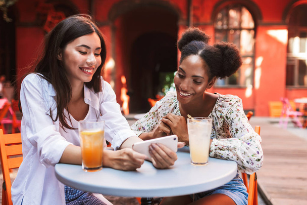Dos hermosas damas felizmente usando el teléfono celular junto con bebidas cerca de la mesa pasar tiempo en el patio de la cafetería
 - Foto, Imagen