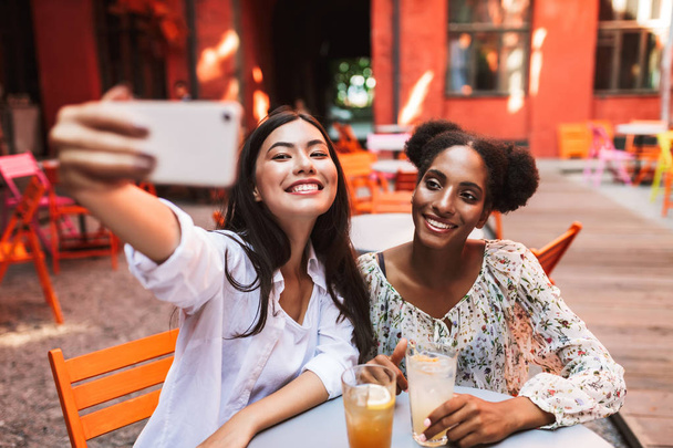 Neşeli kızlar sevinçle birlikte kafe rahat avluda vakit cep telefonu üzerinde fotoğraf çekmek kokteyller ile masada oturan  - Fotoğraf, Görsel