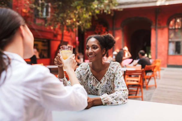 Bardzo uśmiechnięta dziewczyna Afryki w sukienka picia koktajli z przyjacielem radośnie razem spędzać czas w przytulnym courtyard Café - Zdjęcie, obraz