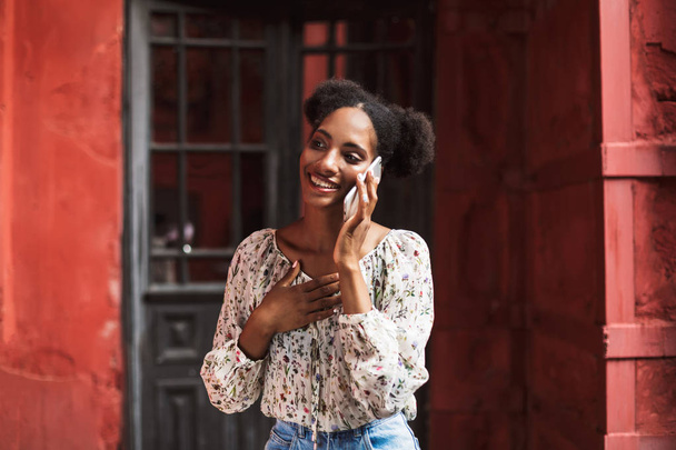 Vrolijke Afrikaans meisje in blouse praten over cellphone gelukkig op zoek opzij buiten - Foto, afbeelding