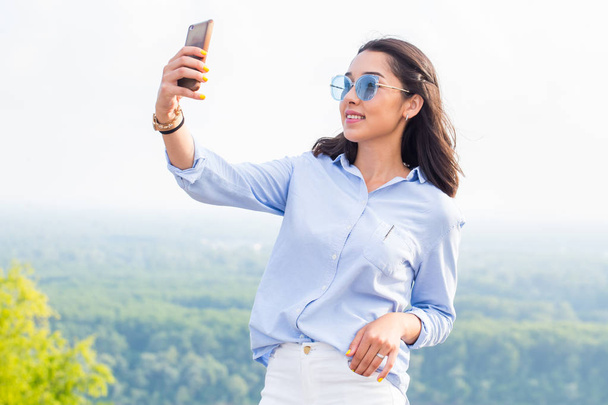 Beautiful smiling woman in blue glasses takes selfies in nature, in the mountains. Modern technology, travel, summer, lifestyle concepts - Foto, afbeelding