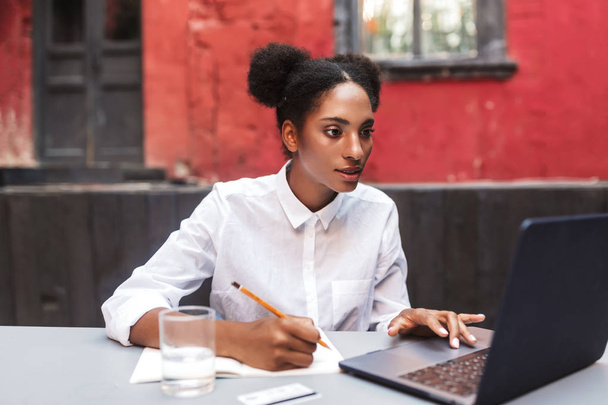 Hermosa chica africana en camisa blanca que trabaja soñadoramente con el ordenador portátil en el patio acogedor de la cafetería
 - Foto, Imagen