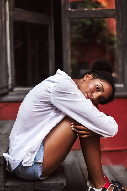 Belle fille africaine aux cheveux bouclés foncés en chemise blanche penchée sur les genoux regardant oniriquement à la caméra avec une vieille fenêtre sur le fond
 - Photo, image