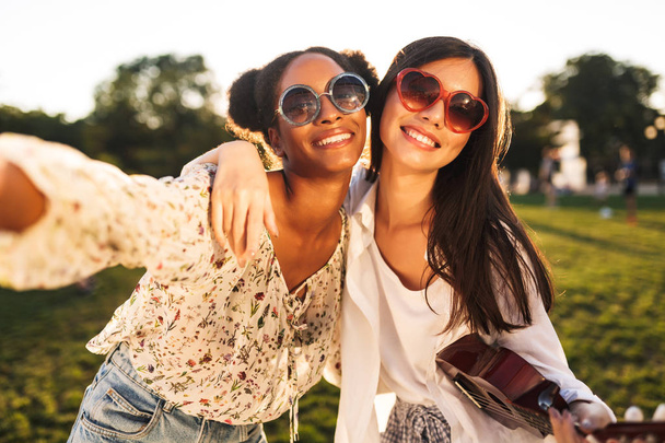 Two beautiful girls in sunglasses friendly hugging each other happily taking photos together in city park - Photo, Image