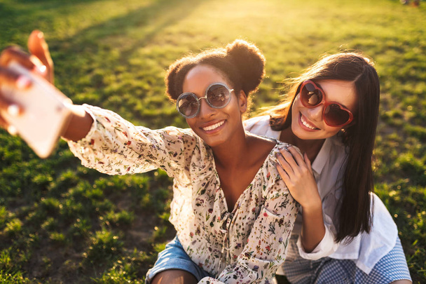 Όμορφα χαμογελαστά κορίτσια σε γυαλιά ηλίου που κάθεται στο γρασίδι χαρωπά λαμβάνοντας φωτογραφίες στο κινητό να ξοδεψουν το χρόνο μαζί στο πάρκο - Φωτογραφία, εικόνα