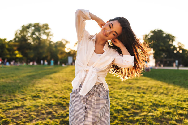 Pretty smiling girl in white shirt joyfully looking aside dancing in city park - Photo, Image