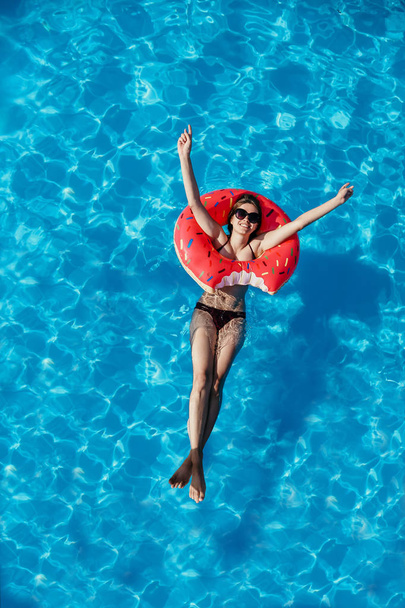 Beautiful young woman with pink donut circle have rest  in blue swimming pool - Foto, imagen