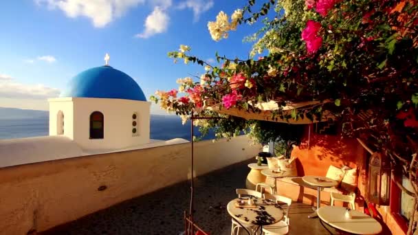Μπλε θόλο εκκλησιών με άνεμο παίζοντας με πολύχρωμο λουλούδι θάμνος βλέπει σε βεράντα από το παραδοσιακό καφενείο στο τυπικό ελληνικό χωριό της Οίας, Σαντορίνη, Ελλάδα. - Πλάνα, βίντεο