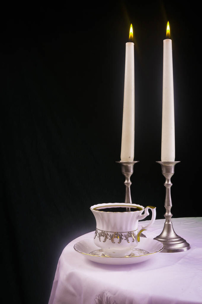 Αναμμένο κερί σε ένα κηροπήγιο με ένα ρετρό φλιτζάνι σε ένα τραπέζι με ένα λευκό πανί - Φωτογραφία, εικόνα