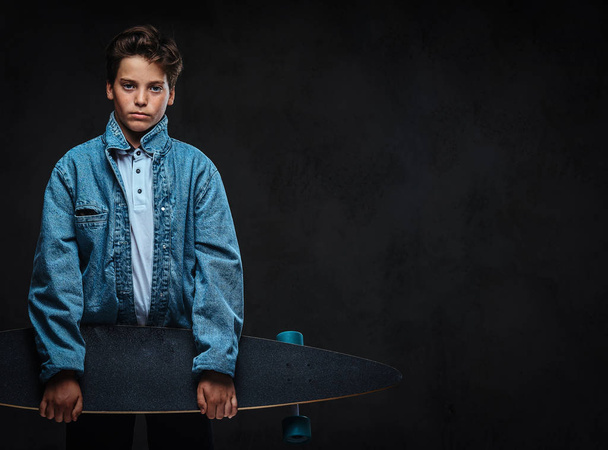 Fashionable young guy dressed in a t-shirt and denim jacket holds a longboard. Isolated on the dark background. - Фото, зображення