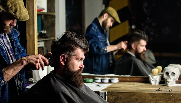 Barber styling hair of bearded client with comb and clipper. Barber with hair clipper works on hairstyle for man with beard, barbershop background. Hipster client getting haircut. Haircut concept - Фото, изображение