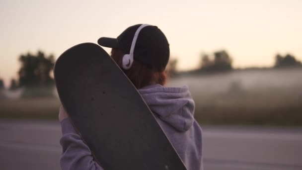 Nahaufnahme eines Hipster-Mädchens mit einem Skateboard in der Hand. geht das rothaarige Mädchen vor dem Hintergrund des Morgennebels die menschenleere Autobahn entlang. Zeitlupe. zurück - Filmmaterial, Video