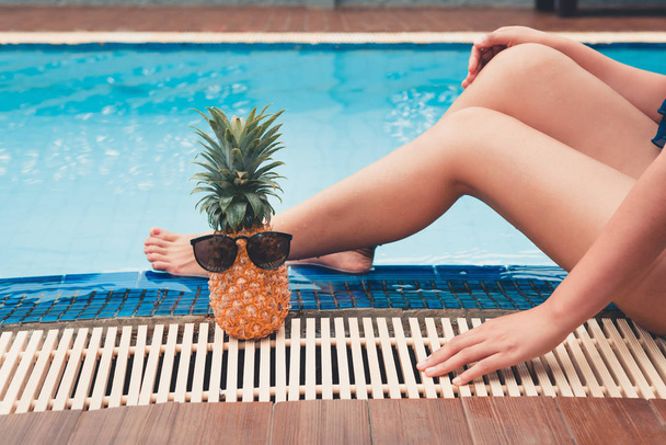 夏のコンセプトは、女性の脚とスイミング プールの横にあるパイナップルをクローズ アップ. - 写真・画像