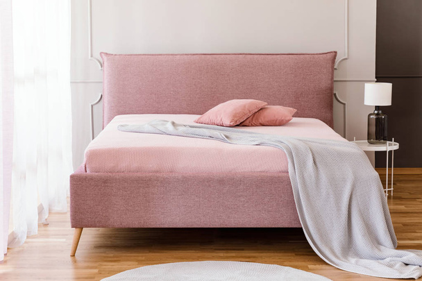 Real photo of pastel pink king-size bed with knit blanket and two pillows standing in bright bedroom interior with molding on the wall - Foto, Imagen