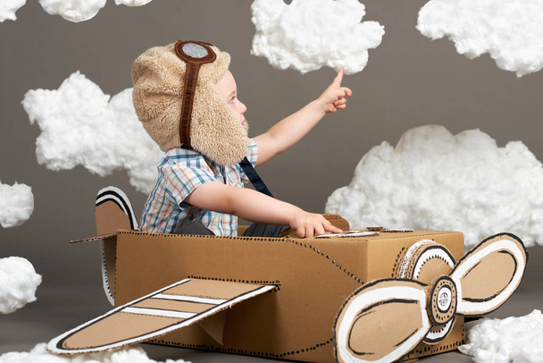 the boy plays in an airplane made of cardboard box and dreams of becoming a pilot, clouds from cotton wool on a gray background, retro style - Photo, Image
