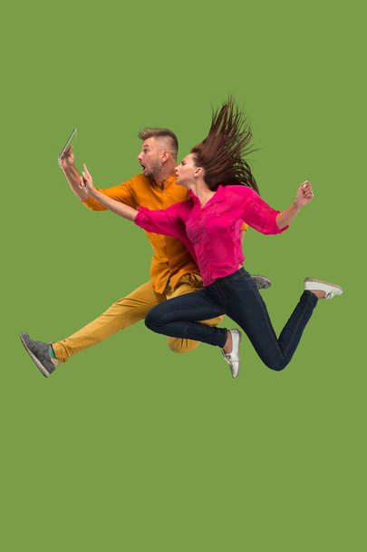 Jump of young couple over green studio background using laptop or tablet gadget while jumping. - Фото, изображение