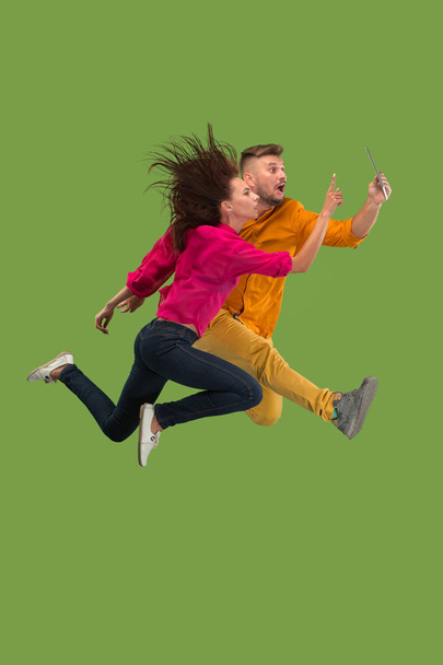 Jump of young couple over green studio background using laptop or tablet gadget while jumping. - Valokuva, kuva