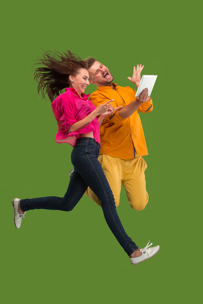 Jump of young couple over red studio background using laptop or tablet gadget while jumping. - Fotografie, Obrázek