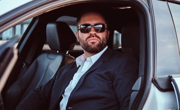 Zakenman in zonnebril gekleed in een formele pak zittend op de voorstoelen in de luxeauto. - Foto, afbeelding