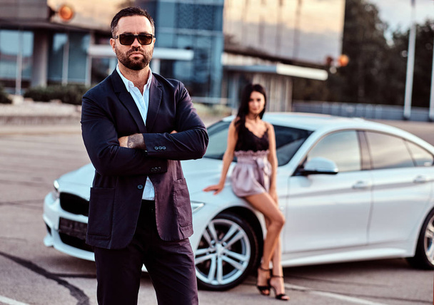 Well-dressed attractive couple leaning on a luxury car outdoors against the skyscraper. - 写真・画像