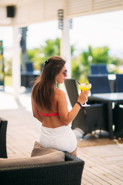 Ελκυστική κοπέλα που στηρίζεται στο beach bar, να πιείτε ένα δροσιστικό κοκτέιλ. Το φωτεινό μαγιό και γυαλιά ηλίου - Φωτογραφία, εικόνα