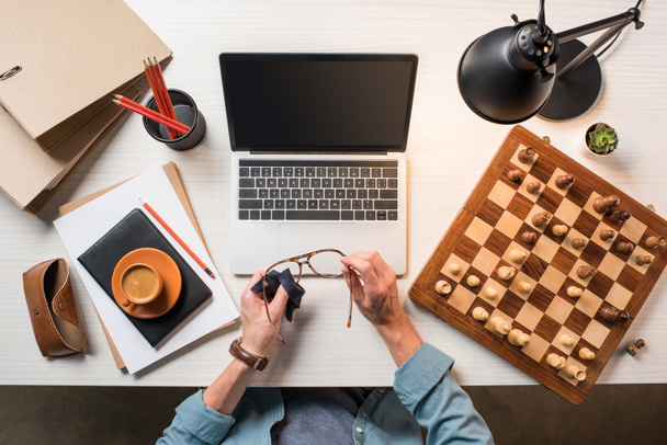 cropped image of male freelancer wiping eyeglasses at working table with laptop, chessboard and coffee  - Foto, Imagen