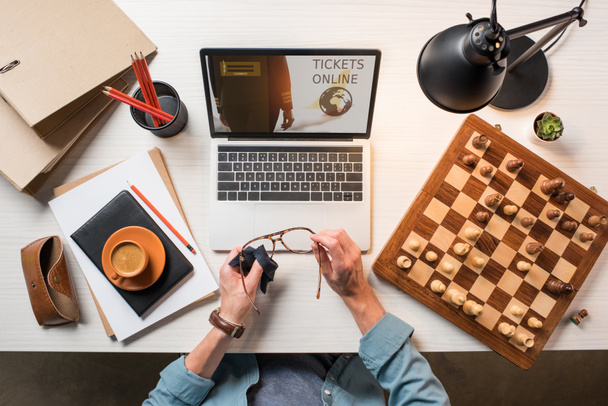 обрезанное изображение мужчины-фрилансера, вытирая очки за столом с шахматной доской, кофе и ноутбуком с билетами онлайн на экране
  - Фото, изображение