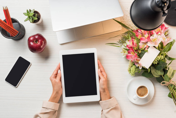 partial view of female freelancer holding digital tablet with blank screen at table with coffee cup, gadgets and flowers with greeting card  - Photo, Image