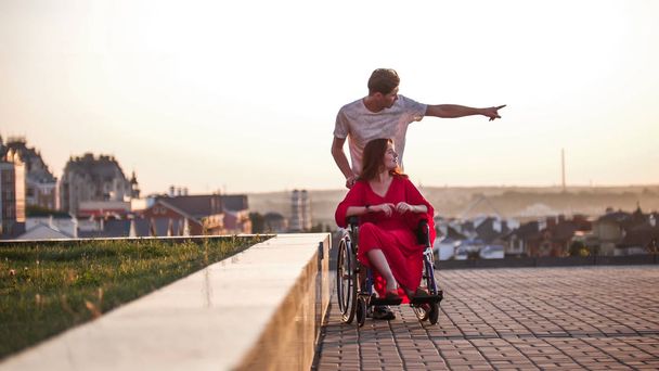 Guy rola sua namorada em uma cadeira de rodas e mostra-lhe algo com a mão na distância, de perto
 - Foto, Imagem